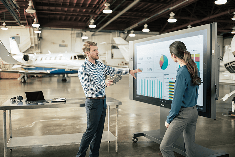 Deux personnes travaillent dans un hangar avec un avion. Ils travaillent sur un Surface Hub qui montre des graphiques Power BI.