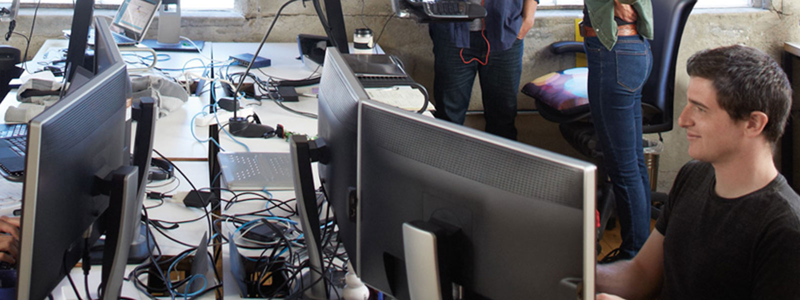 Un espace de travail encombré avec plusieurs écrans d'ordinateur, des câbles et une personne travaillant devant deux écrans. A l'arrière-plan, il y a d'autres personnes, mais elles ne sont pas clairement visibles en raison de la focalisation sur le premier plan.